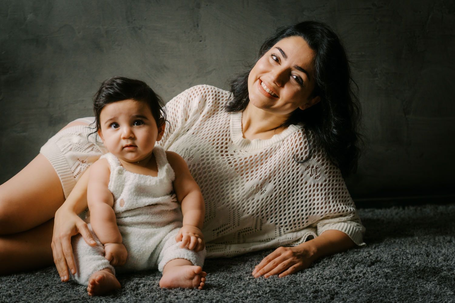 A young mother in a cream knit sweater lies on a gray carpet smiling beside her cute baby in a white romper.