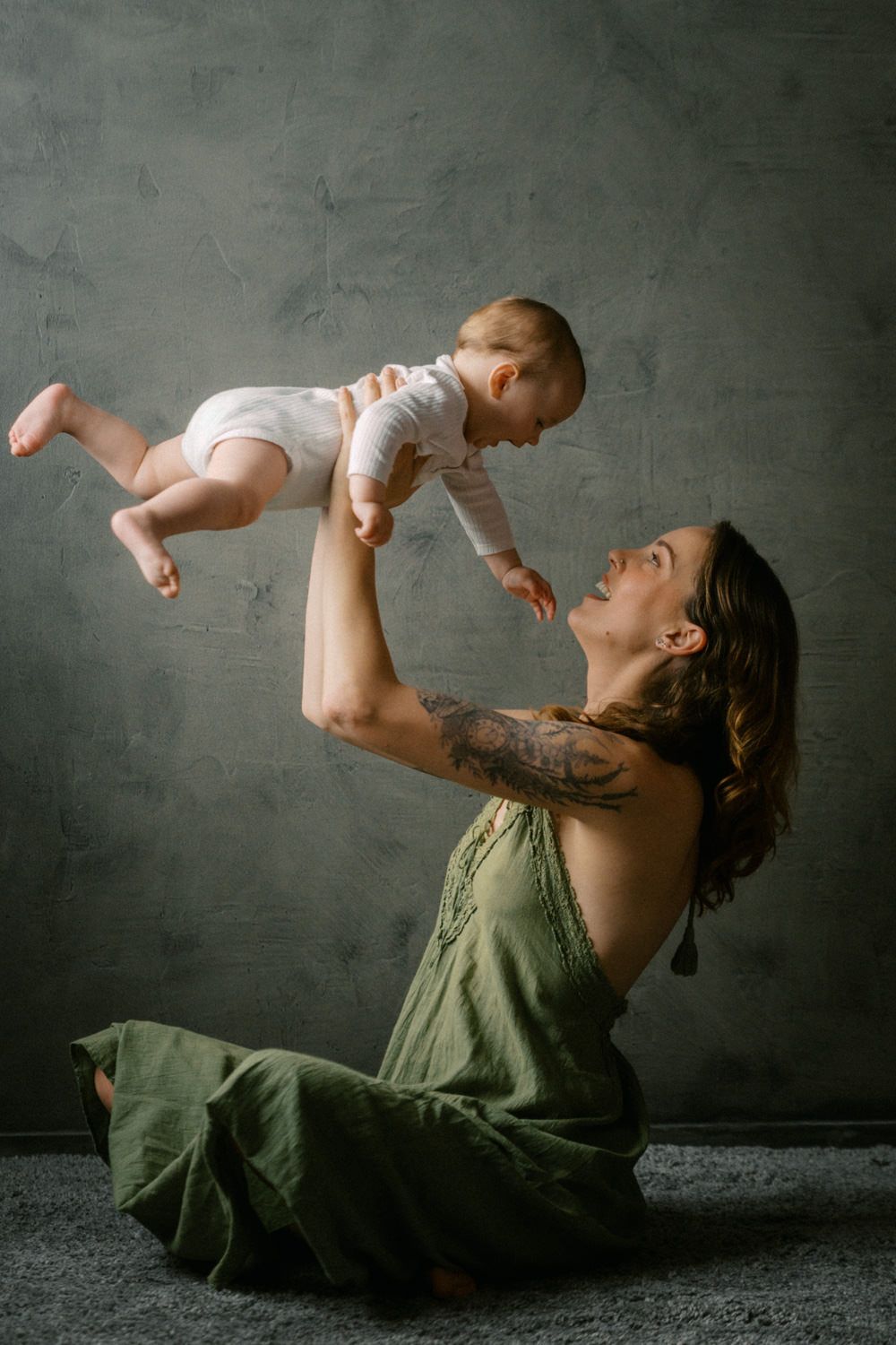 A tattooed mother in a green dress sits on the floor, joyfully lifting her baby in the air indoors.