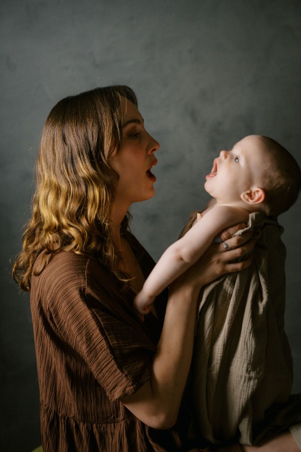 A mother in a brown dress holds her laughing baby up in the air, sharing a joyful moment against a gray background.