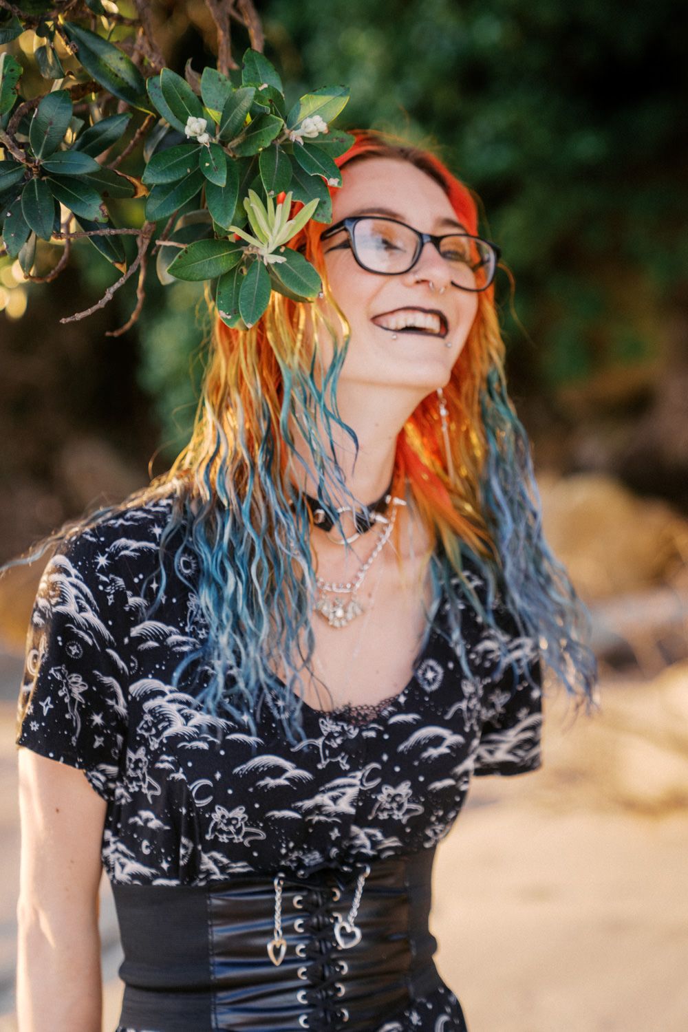 Smiling woman with colorful red and blue dyed hair, black glasses, wearing a dark patterned dress outdoors.
