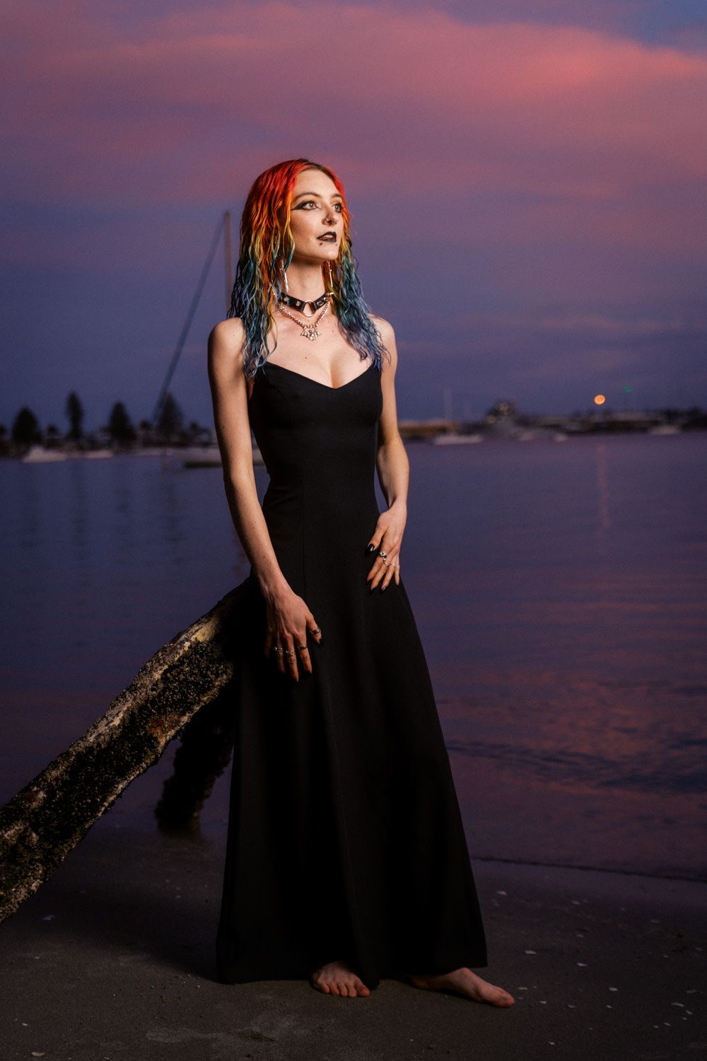Woman in black gown with colorful dreadlocks poses barefoot by waterfront at dramatic purple sunset.