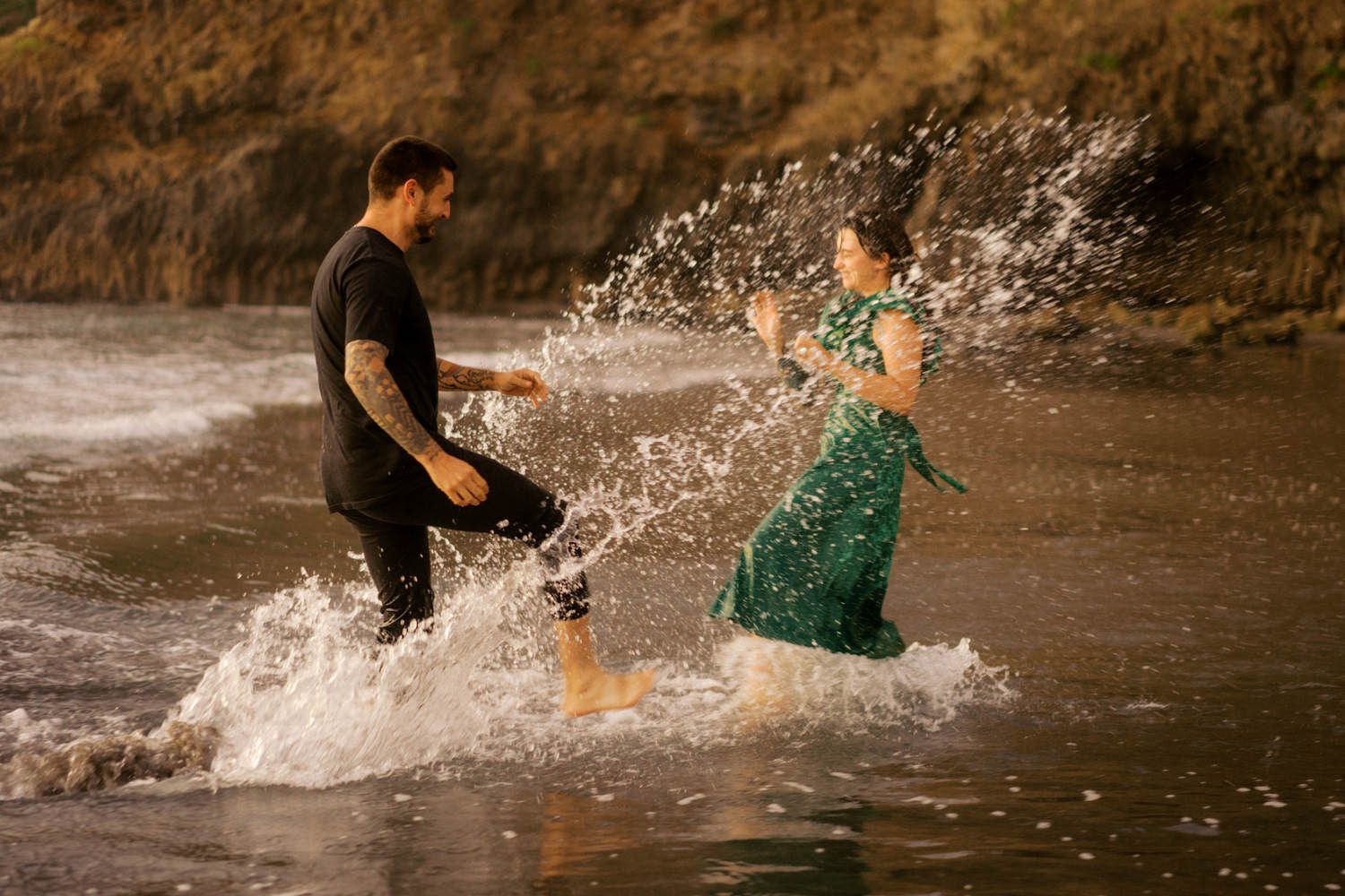 Couple splashing in ocean waves at beach, man in black wetsuit and woman in green dress playing in water.
