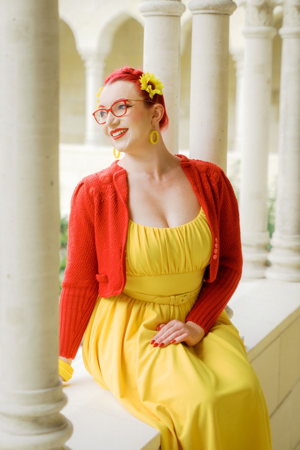 Woman in yellow dress and red cardigan with matching red glasses and hair, yellow floral earrings, sitting among white columns.