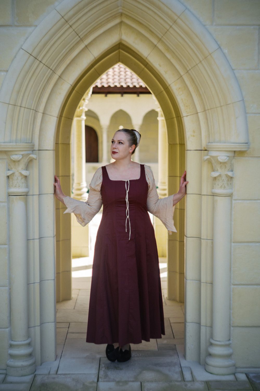 Woman in medieval burgundy dress poses under Gothic stone arch in a sunlit courtyard.