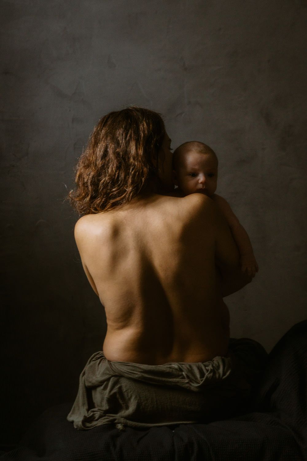 A topless mother holds her newborn baby against her bare back in a moody, dark studio portrait.