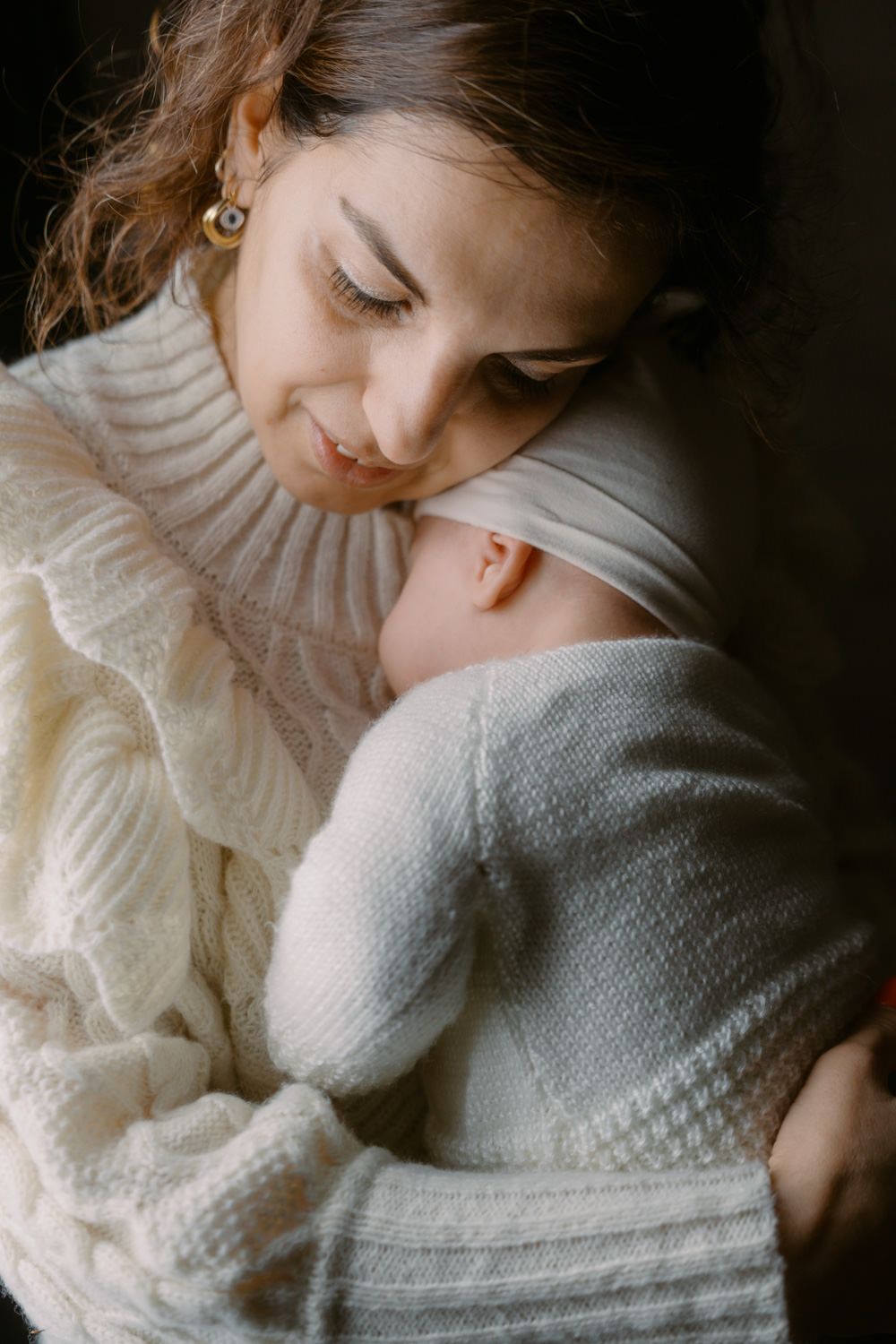 A mother in a white knit sweater tenderly holds her newborn baby, both dressed in soft white, sharing a peaceful moment.