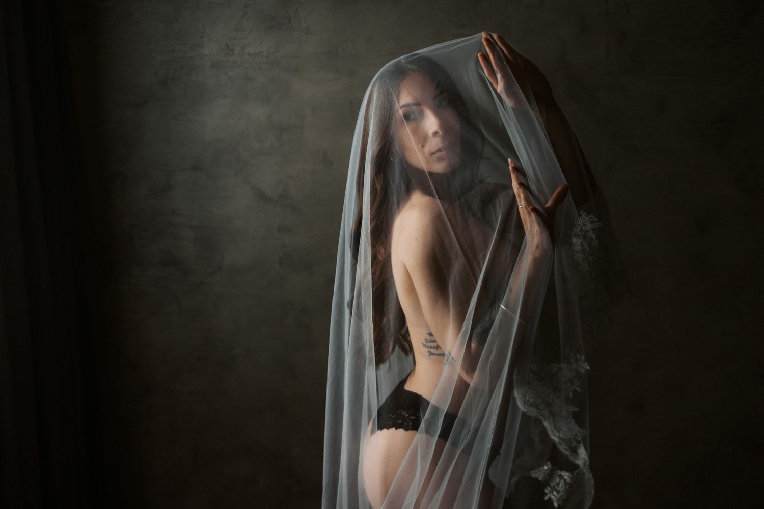 A woman draped in a sheer veil poses in black lingerie against a dark textured studio background.