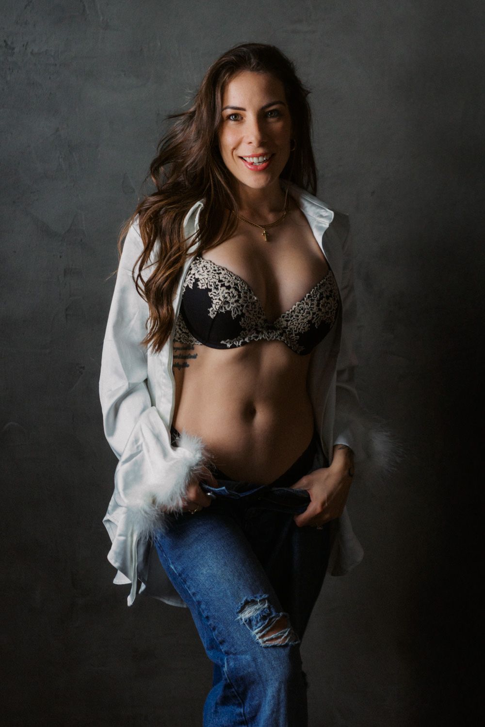 Woman in black lace bra, open white shirt, and ripped jeans posing confidently against a dark studio background.