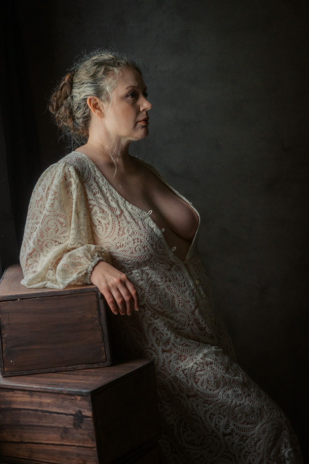 Woman in elegant lace dress seated on wooden crate in moody studio portrait photography.