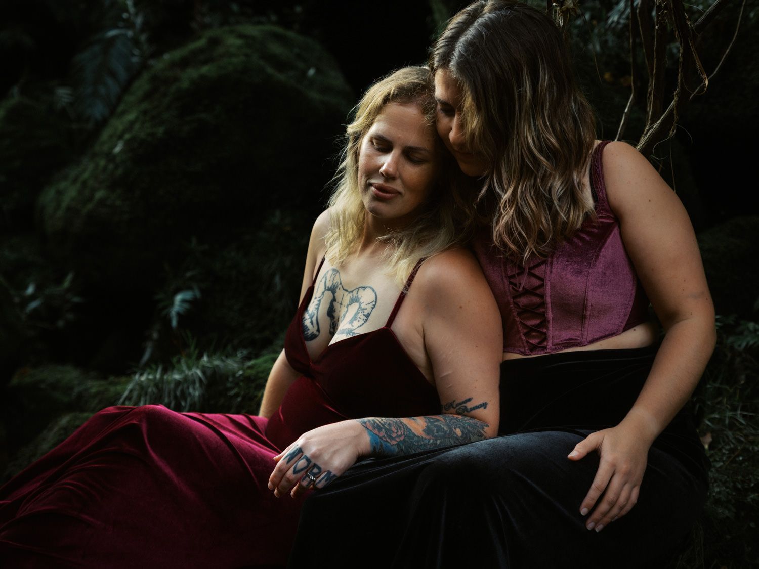 Two women in elegant gowns share an intimate moment in a lush forest setting, adorned with tattoos.
