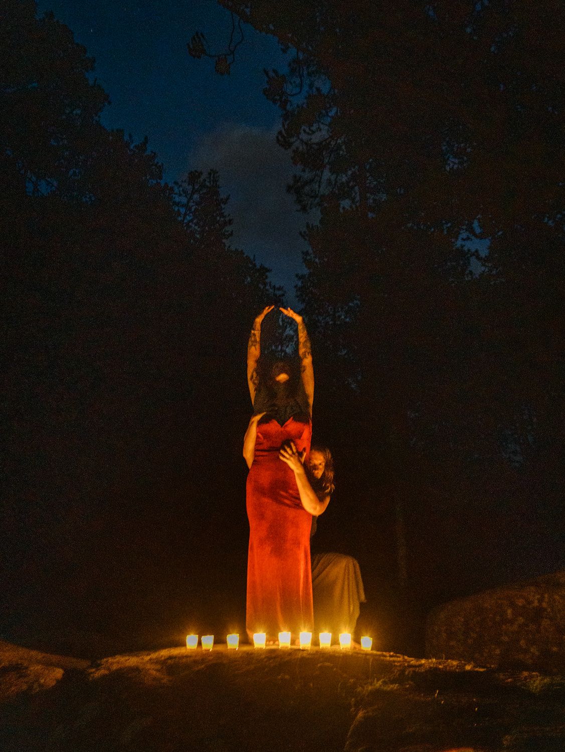 Person in red dress kneeling before candles at night in forest with arms raised dramatically.