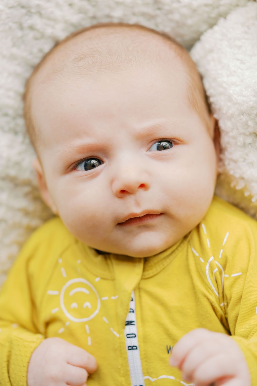 Newborn baby with blue eyes wearing a yellow sunshine onesie lying on a soft white blanket.