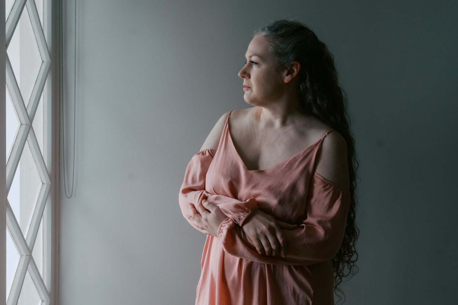 Woman in pink satin dress gazing thoughtfully out a window, with long curly hair in a serene indoor setting.