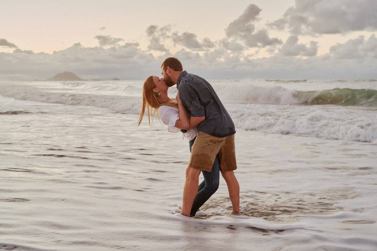 Couple sharing a romantic kiss on a sandy beach with ocean waves crashing around their feet at sunset.