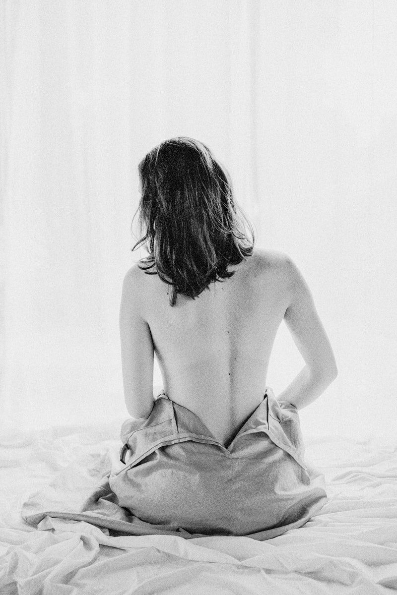 Black and white photo of a woman sitting on a bed, viewed from behind with bare back and draped fabric.