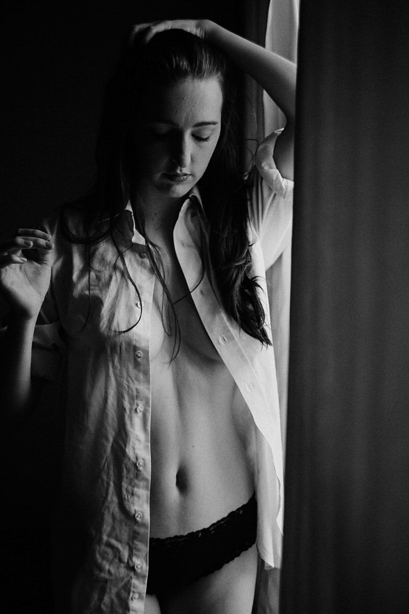 Black and white boudoir photo of a young woman in an open white shirt and dark underwear posing by a curtain.