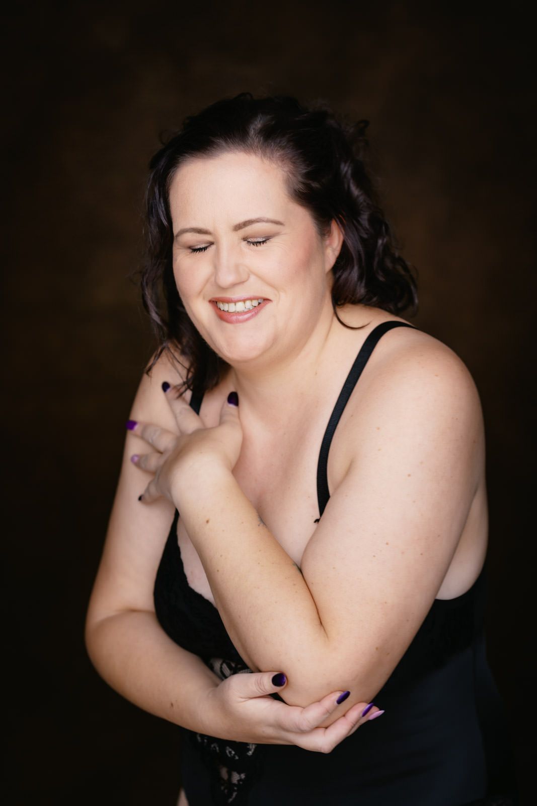 A smiling plus-size woman in a black dress poses confidently against a dark brown studio background.