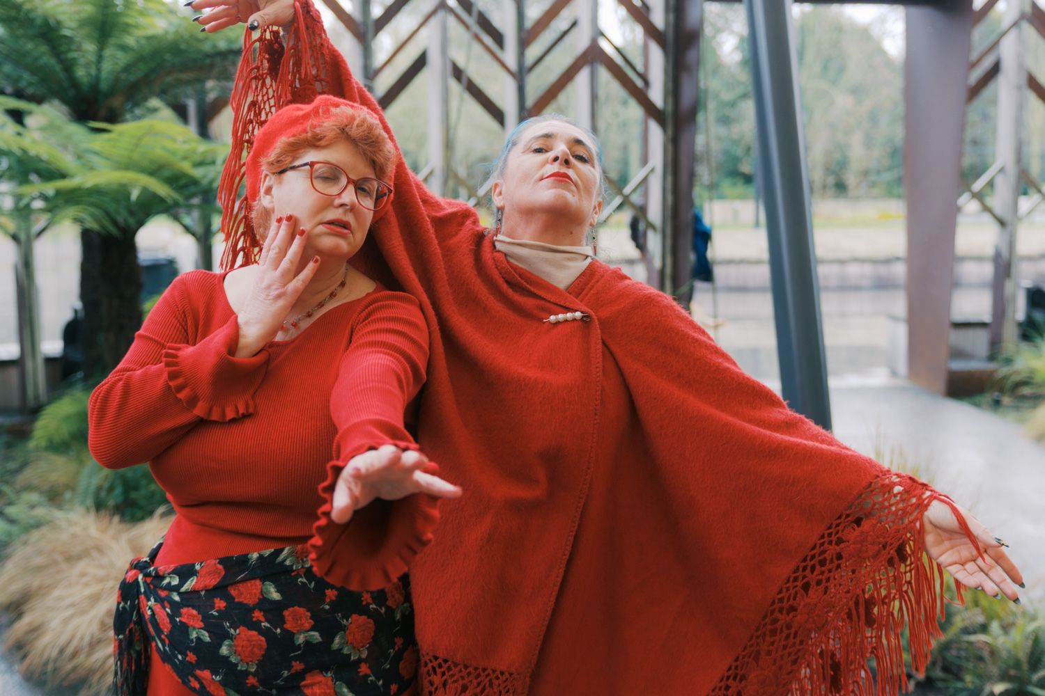 Two women in red outfits pose dramatically indoors, one wearing a fringed shawl and glasses, with green plants behind.