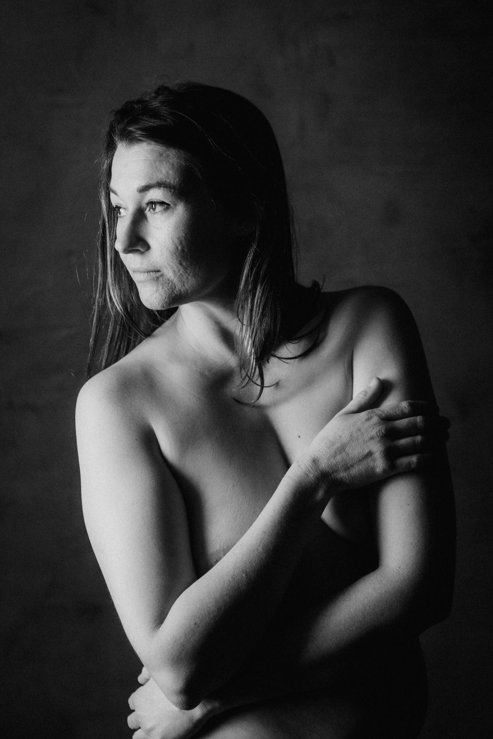 Black and white artistic portrait of a woman with long hair crossing her arms, gazing away in a moody studio setting.