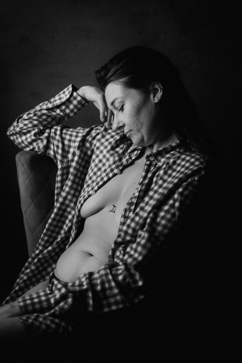 Black and white portrait of a woman in an open plaid shirt seated in a chair, eyes closed, in dramatic lighting.