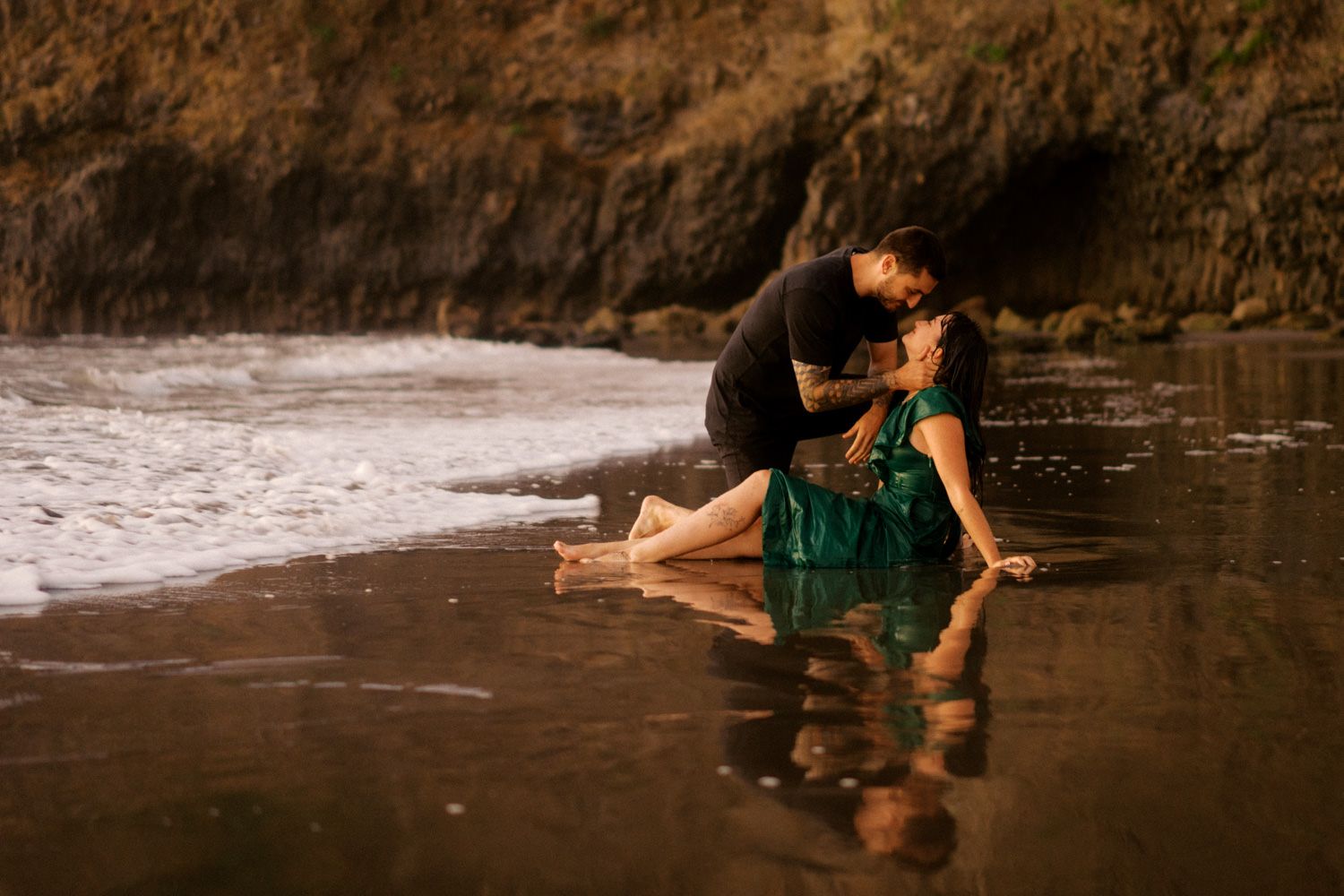 Couple sharing a romantic kiss on a dark sandy beach, woman in green dress sitting in shallow ocean waves.
