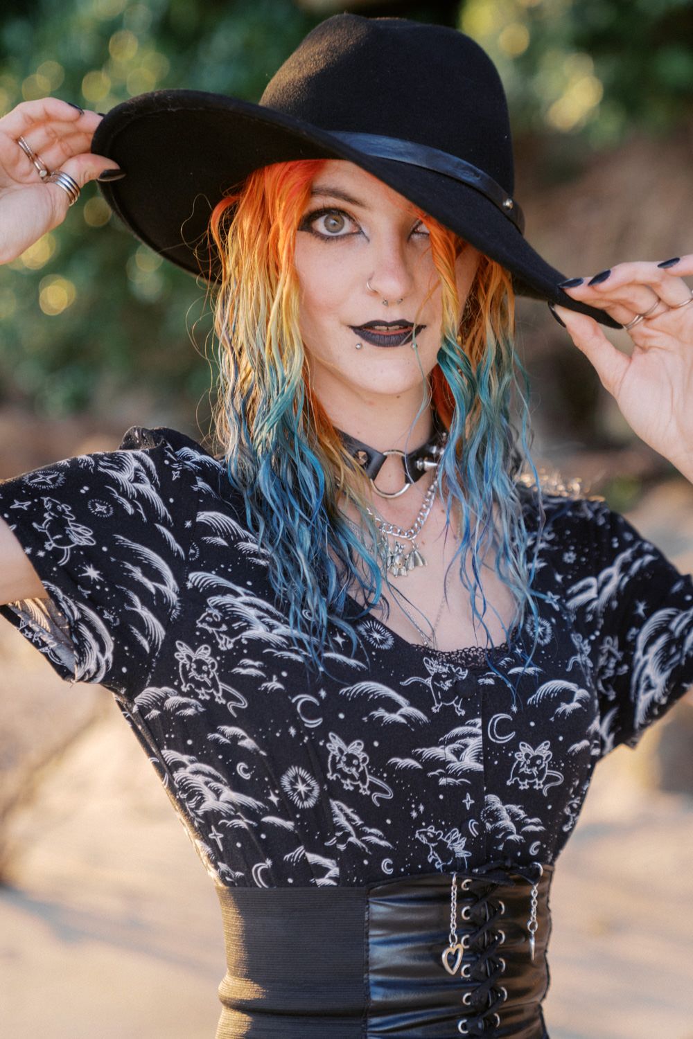 Woman with colorful orange and blue hair wearing black witch hat, dark lipstick, and gothic celestial print top.