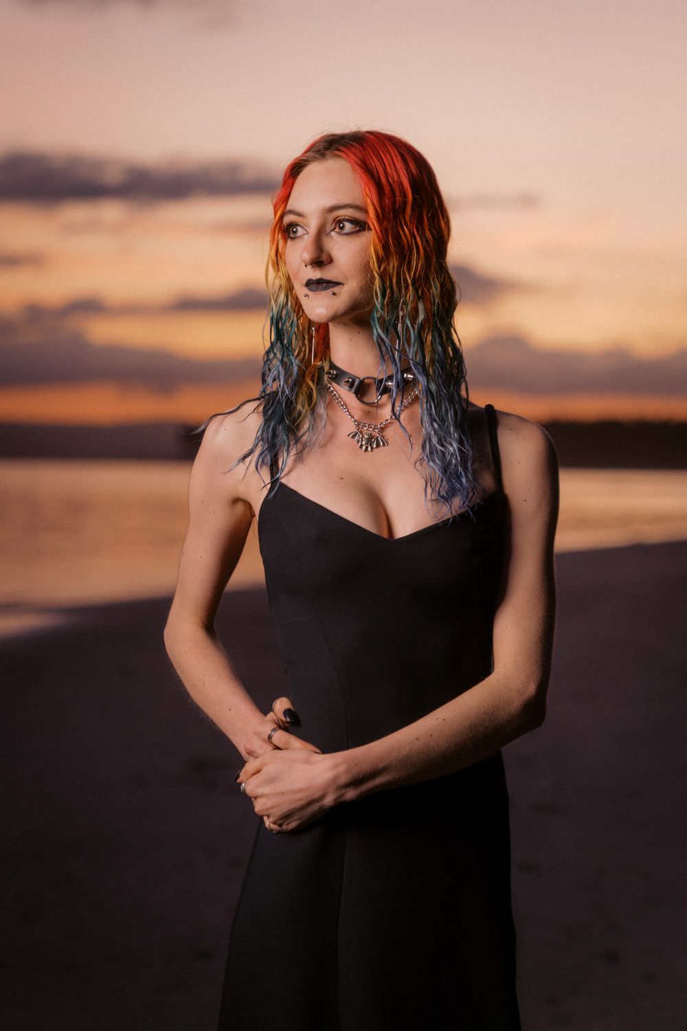 Woman with colorful red and blue hair in black dress poses on beach at sunset wearing statement necklace.