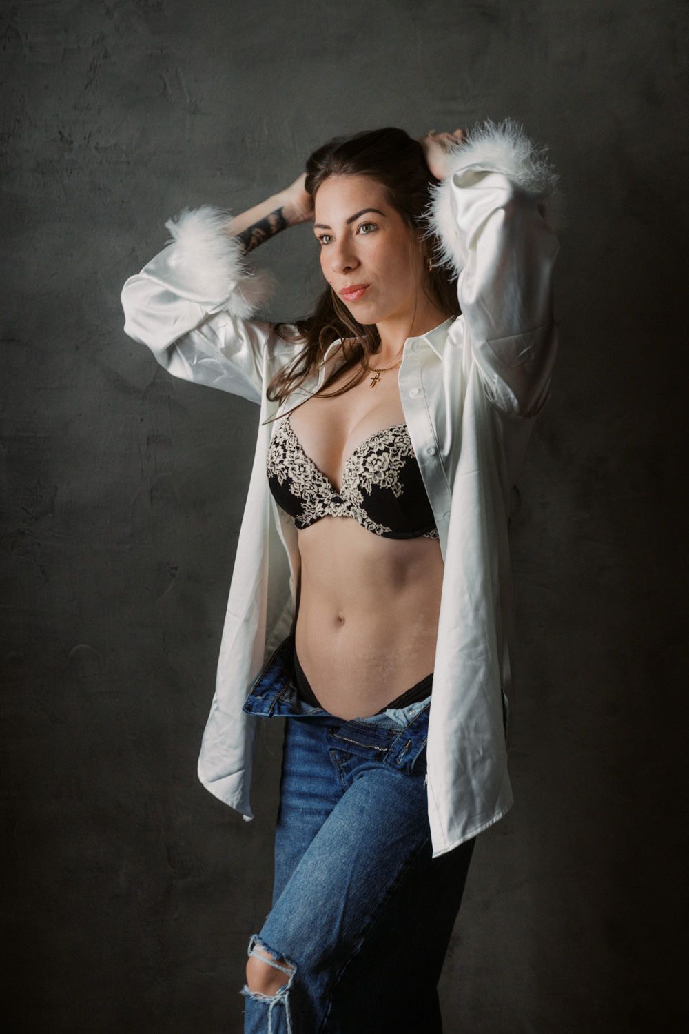 Woman in open white feathered shirt, black embellished bra, and ripped jeans posing against gray wall.