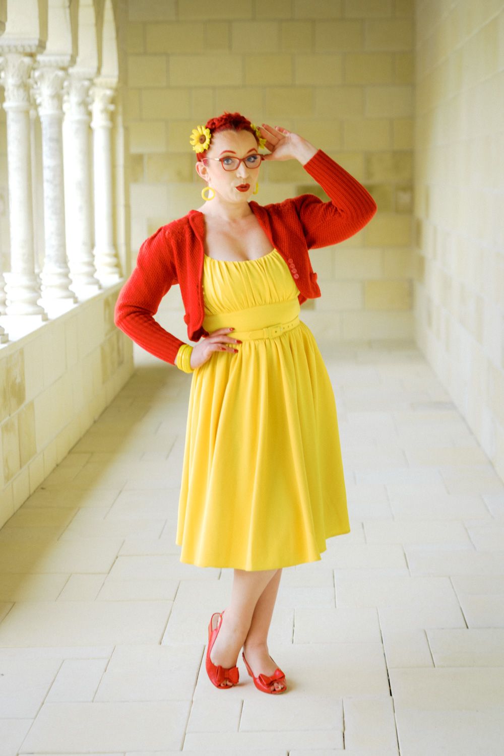 Woman in yellow sundress and red cardigan with matching heels poses in elegant white columned hallway.