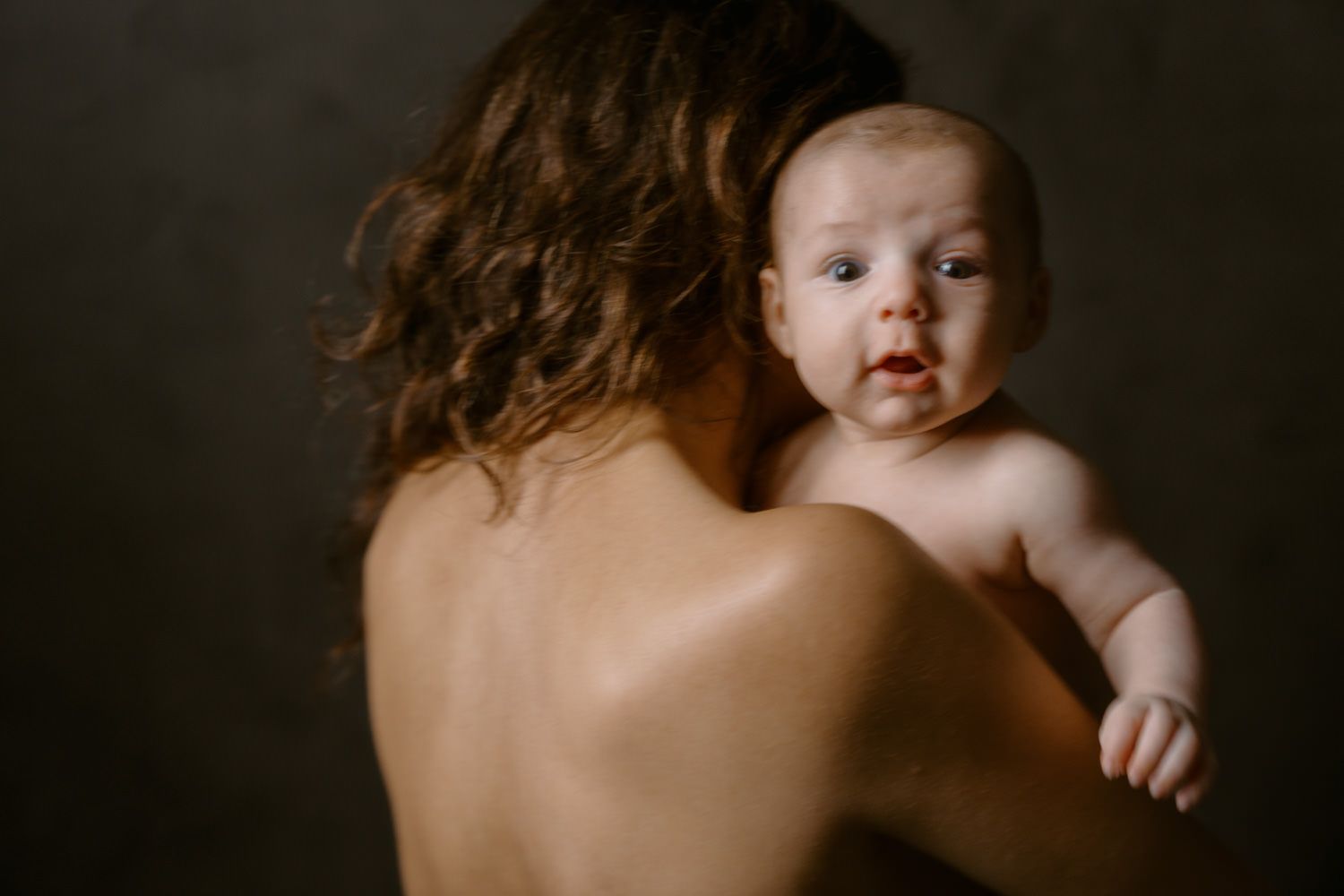 A mother holds her naked newborn baby against her bare shoulder in a tender, artistic portrait with dark background.