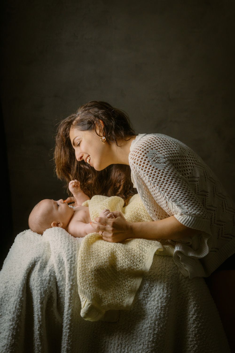 Mother in white knit dress tenderly leaning over newborn baby wrapped in yellow blanket on dark background.