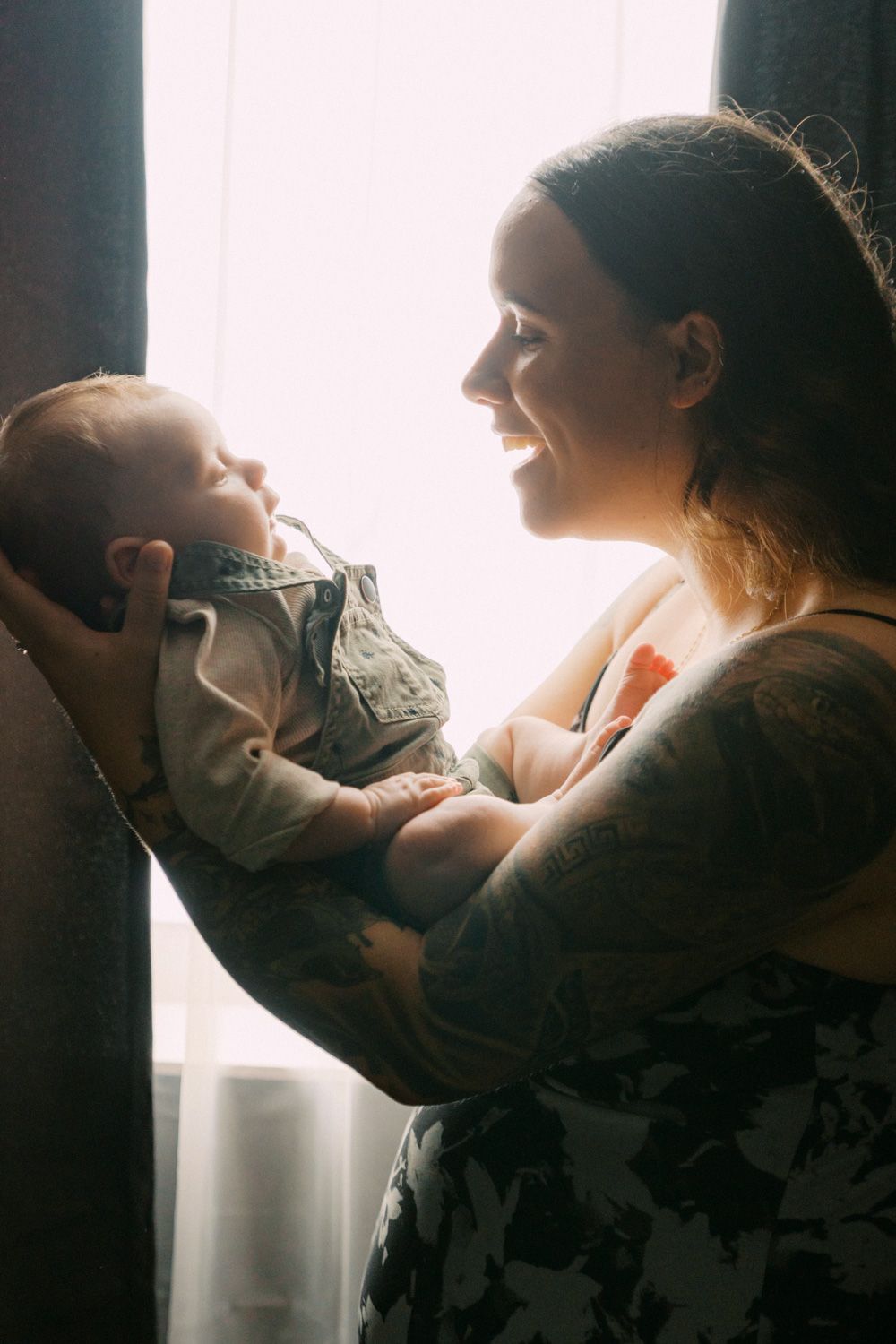 Tattooed mother smiling and holding newborn baby near a bright window in a tender moment.