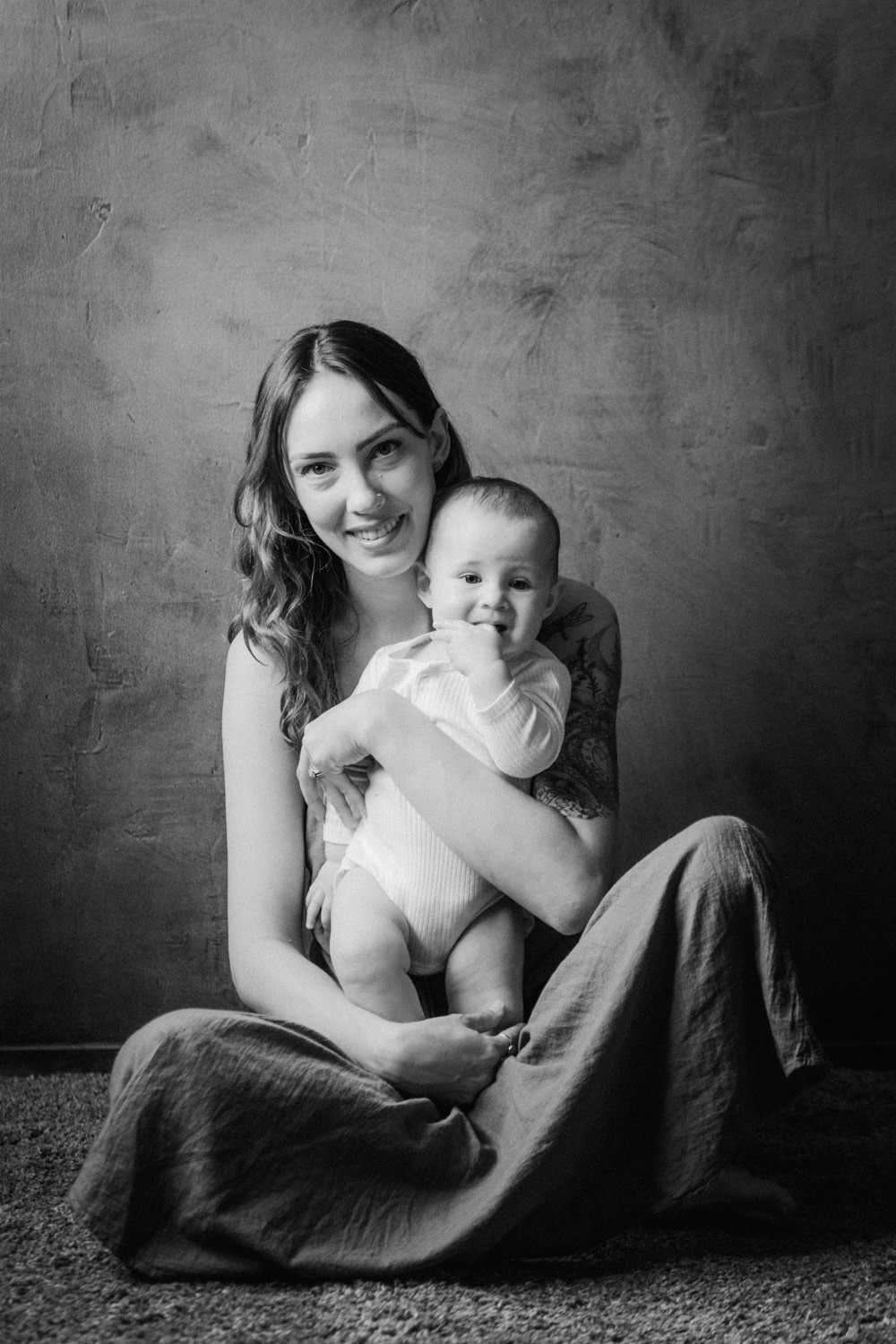 Black and white portrait of a smiling mother sitting on the floor holding her happy baby in a studio setting.