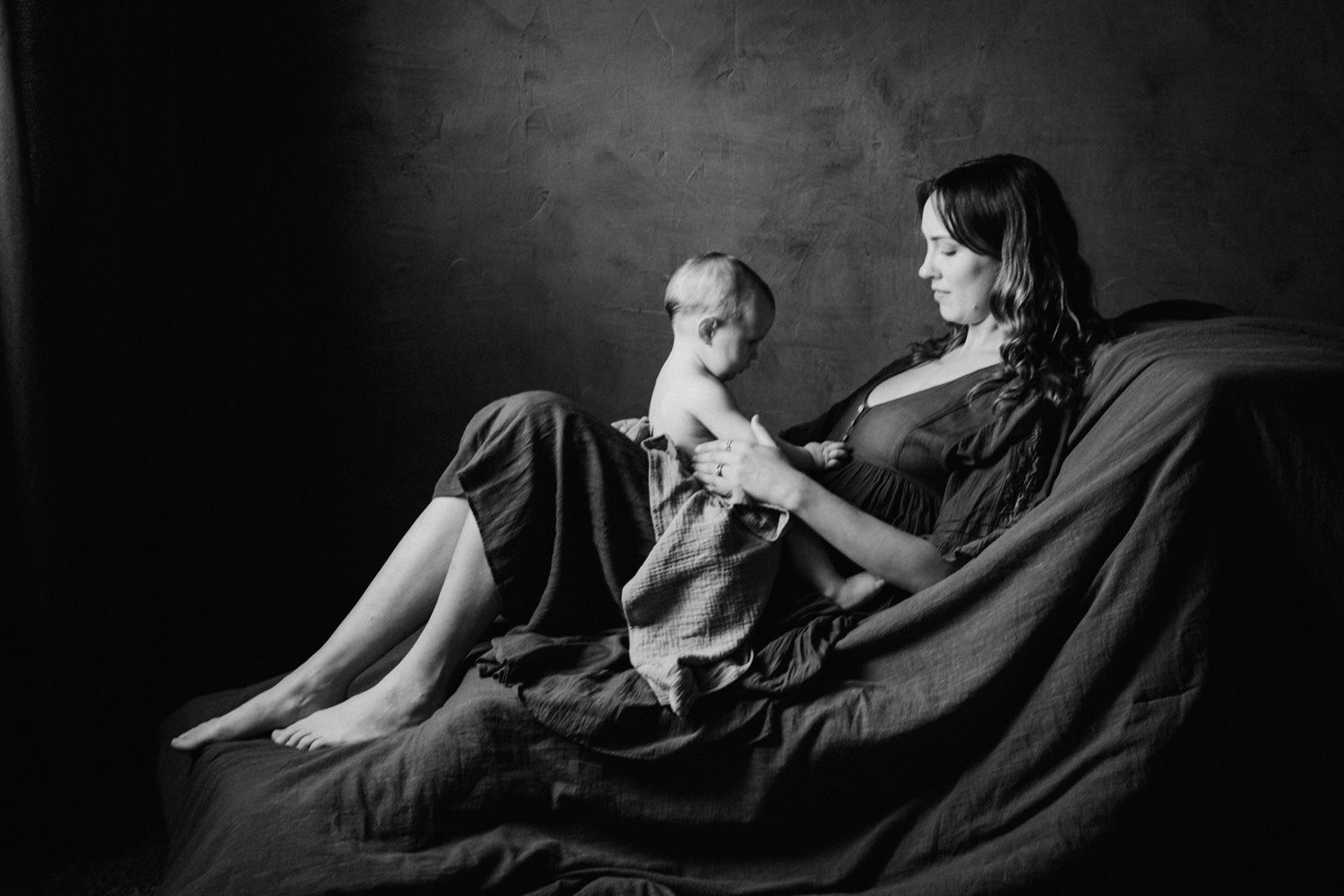 Black and white photo of a mother and baby sharing a tender moment on a draped couch in a studio.