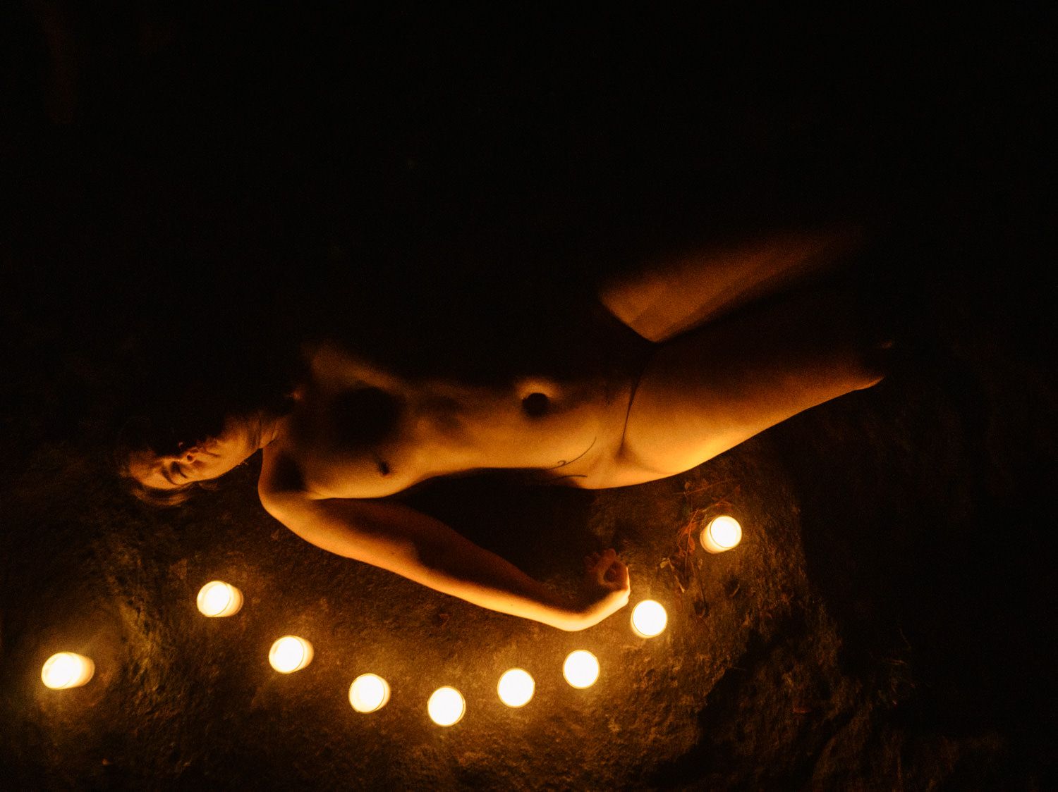 A nude figure reclining on a rock surrounded by glowing candles in dramatic candlelight photography.