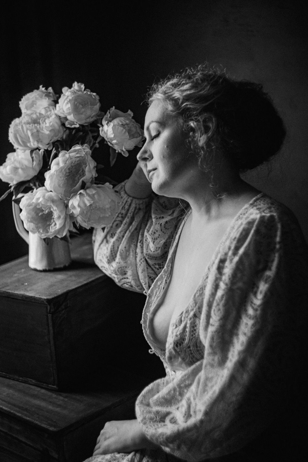 Black and white portrait of a woman in lace robe smelling white peonies in a vase, eyes closed peacefully.