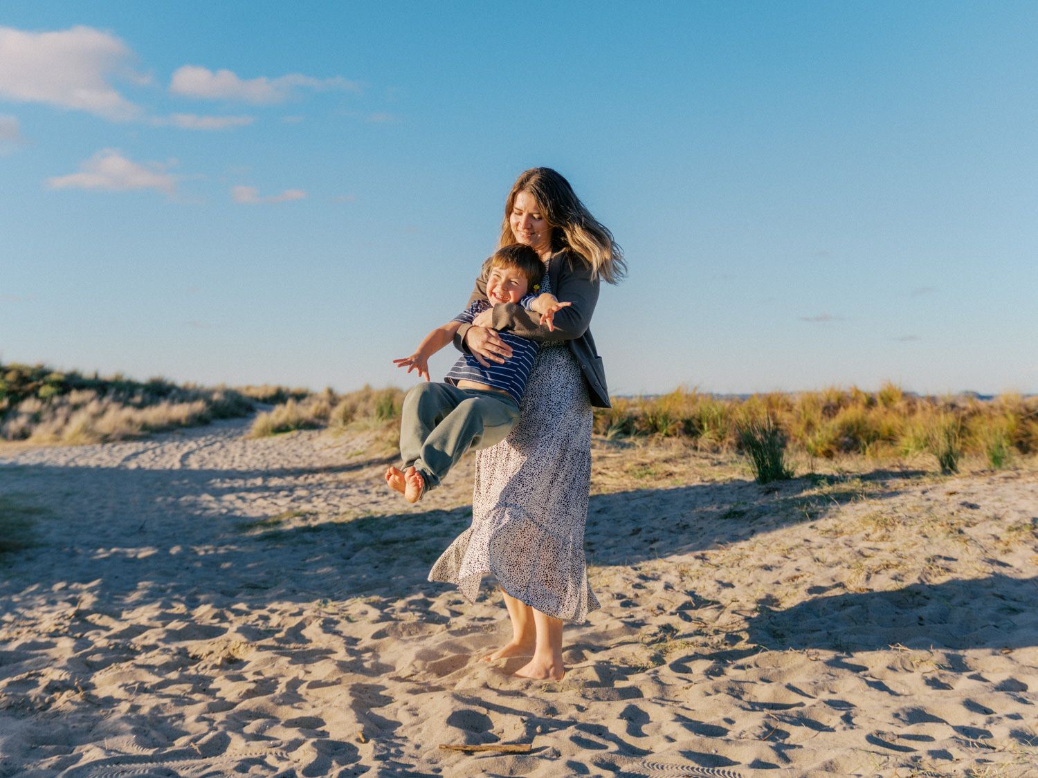 A woman carries a laughing child on her back while playing on a sunny sandy beach with dunes and blue sky.