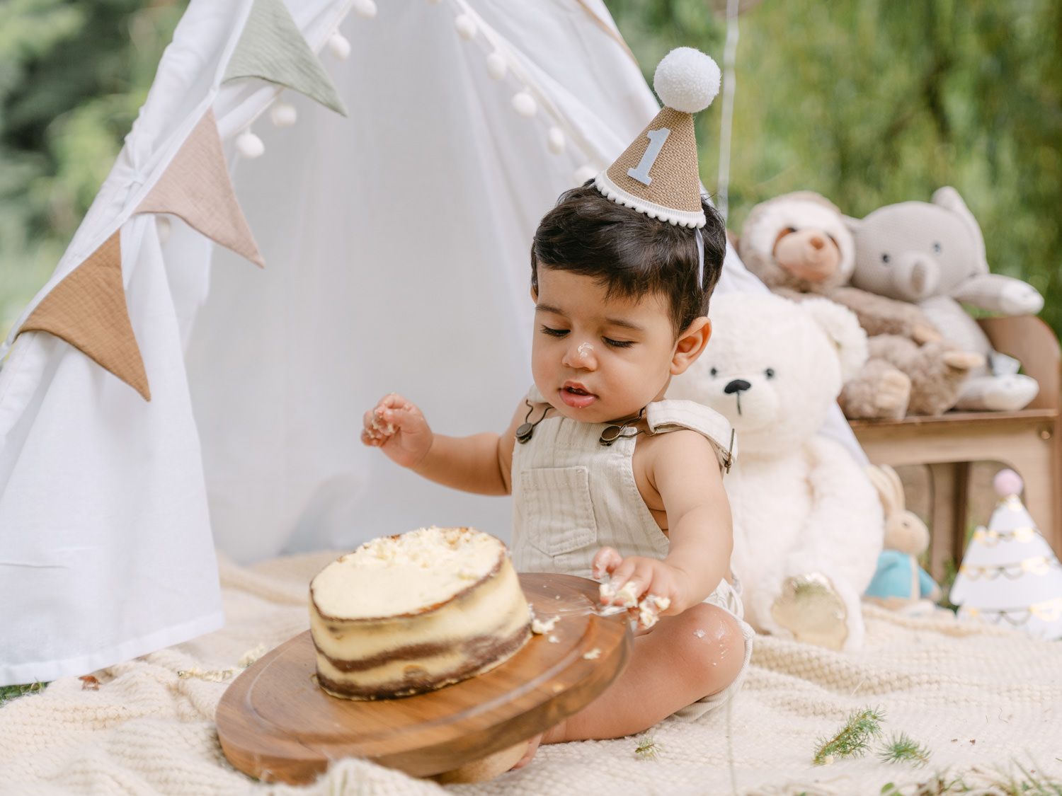 Baby in a party hat smashes first birthday cake outdoors near a teepee with teddy bears in a neutral boho setting.