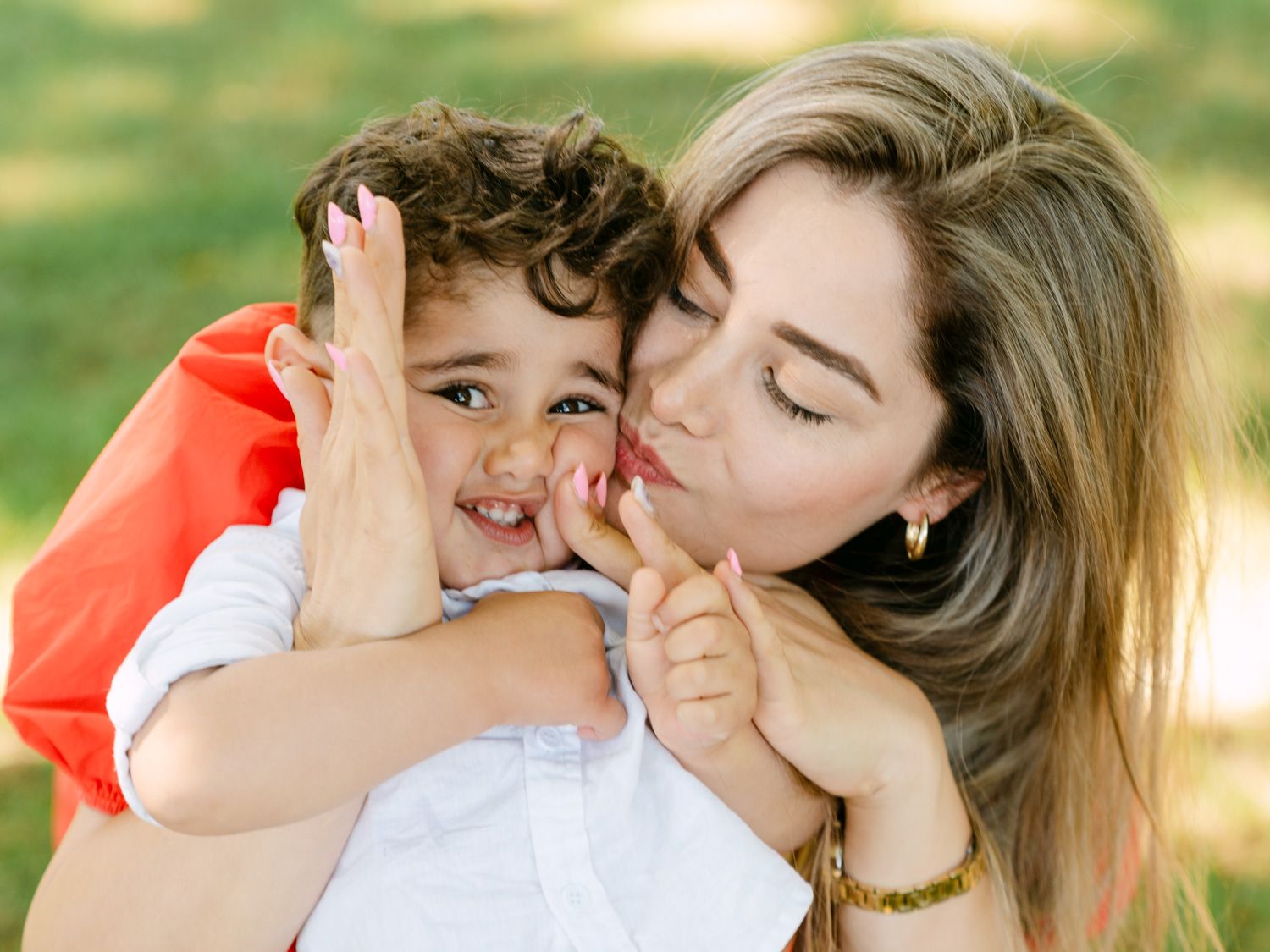 A loving mother kisses her smiling toddler boy outdoors in a sunny green park setting.