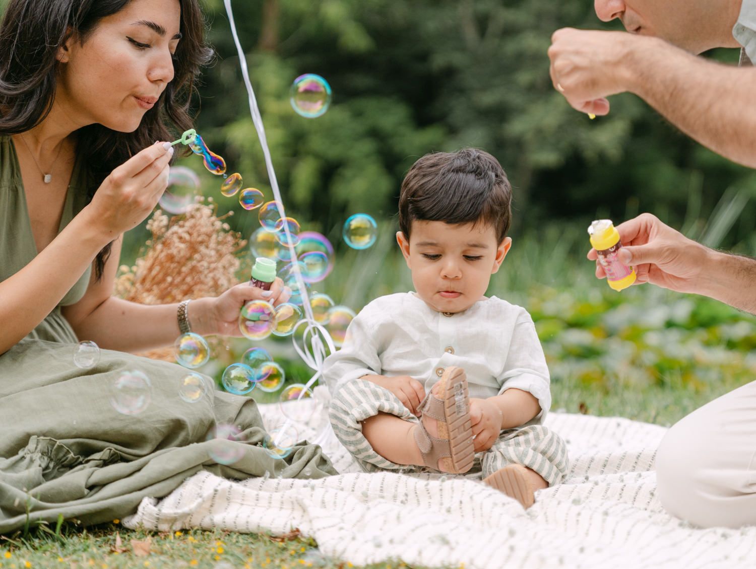 Family blowing bubbles with toddler on picnic blanket outdoors in lush green garden setting.