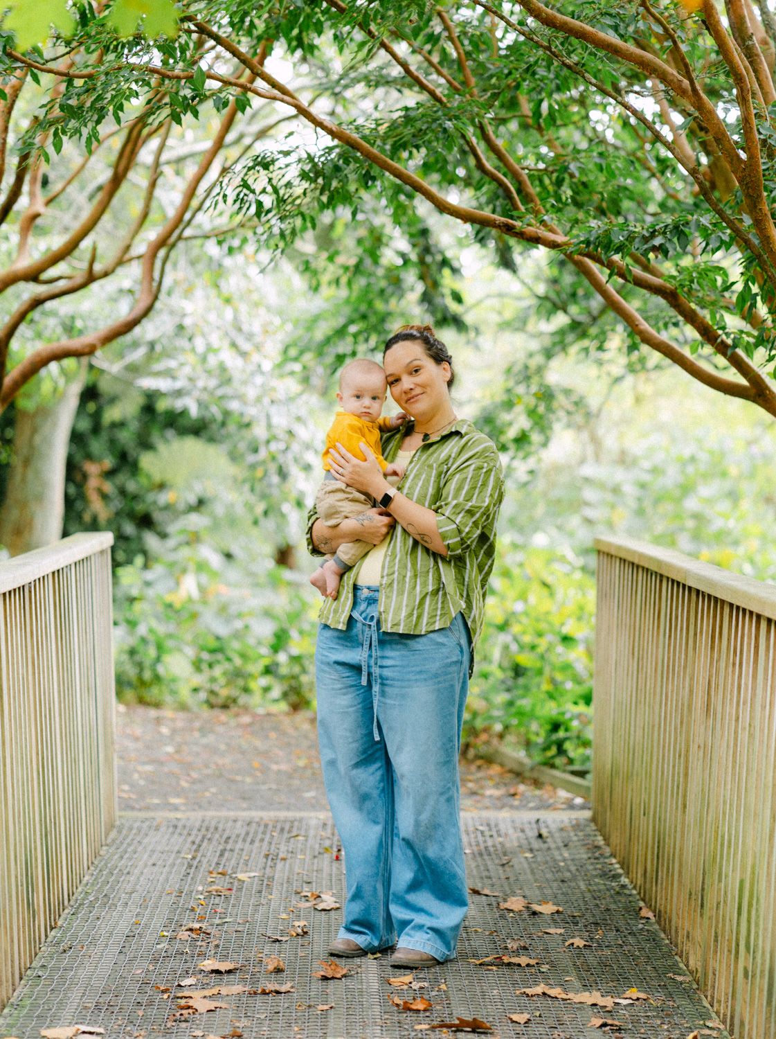 Mother holding baby on wooden bridge surrounded by lush green trees in a park.