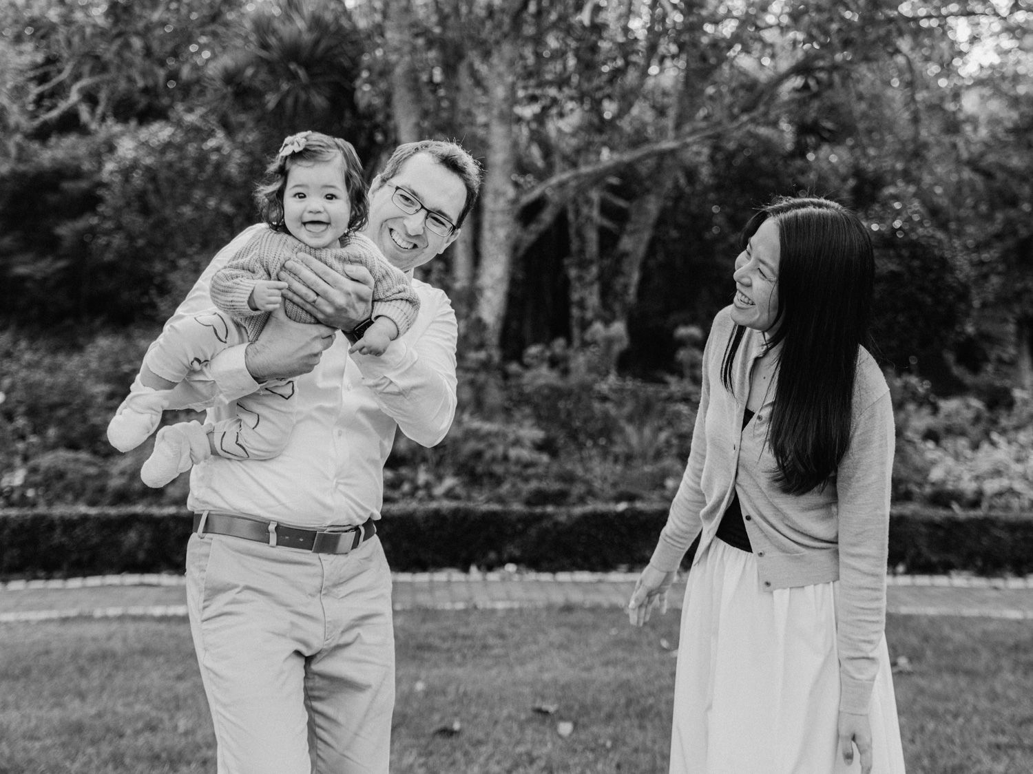 Black and white photo of happy family with father lifting baby while mother smiles in garden.