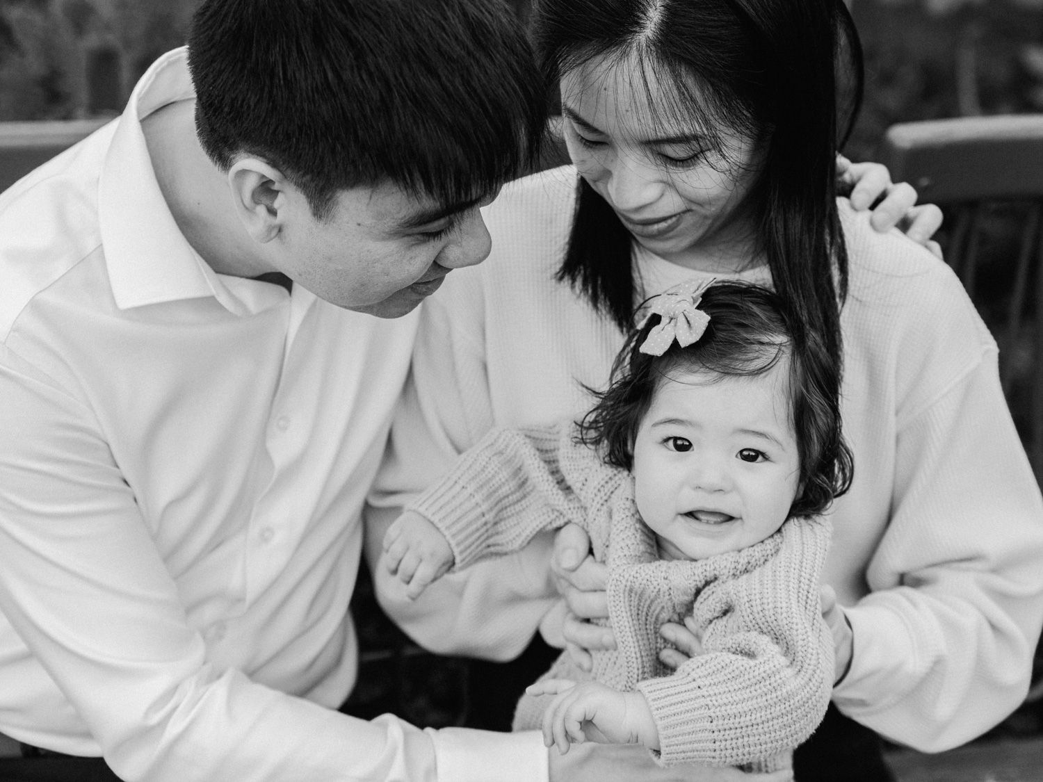 Black and white family portrait of parents lovingly gazing at their smiling toddler in a cozy knit sweater.