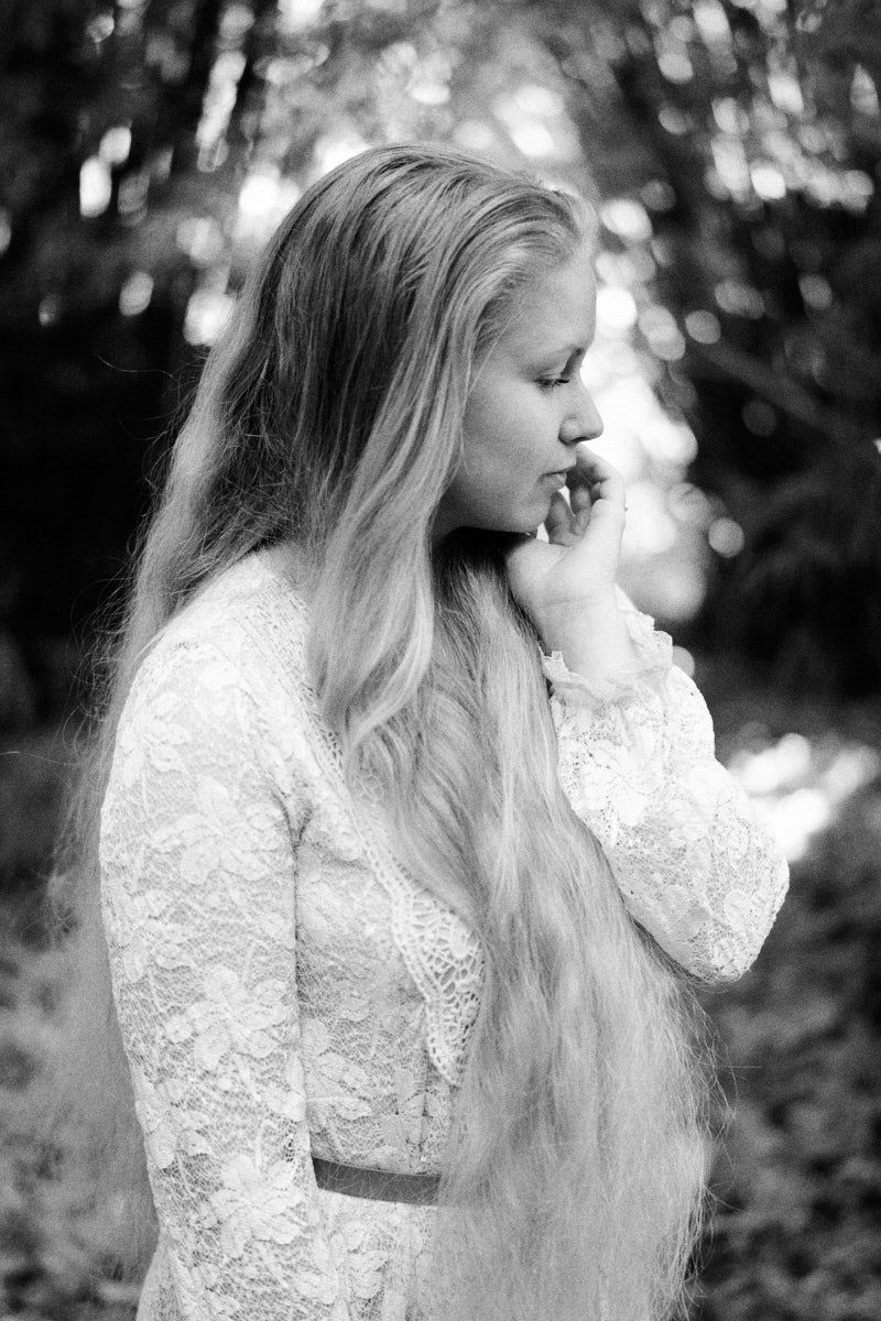 Black and white portrait of a young blonde woman in lace dress, looking down thoughtfully in a forest setting.