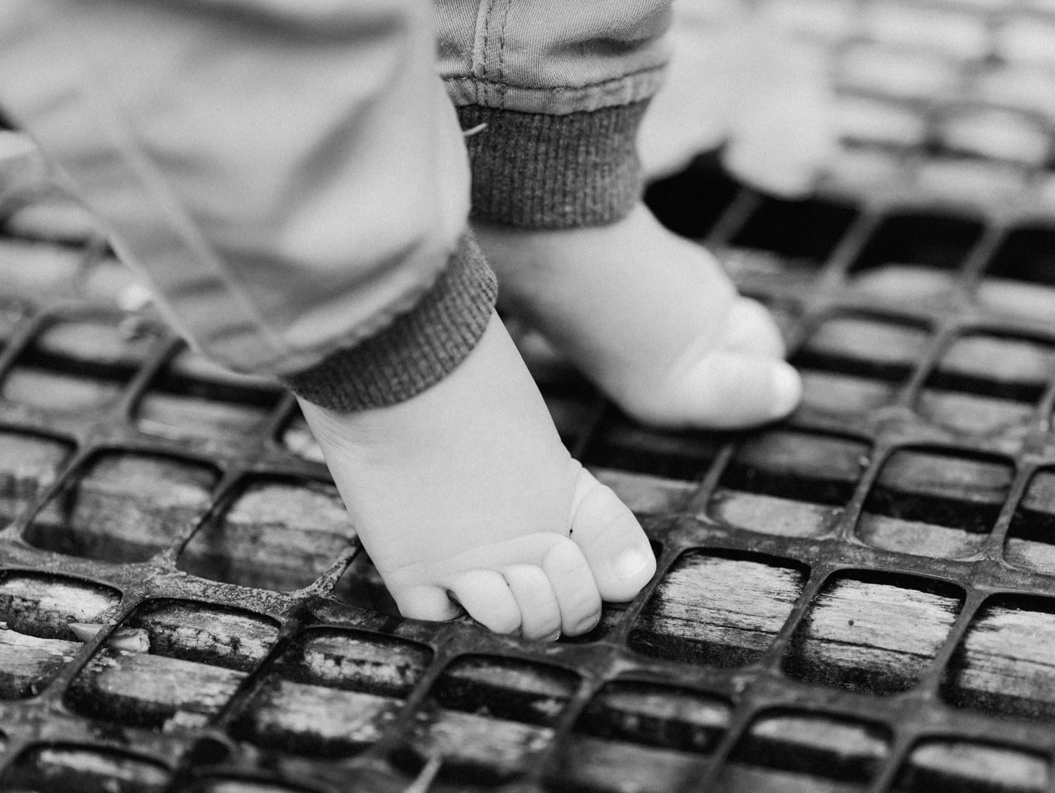 Close-up black and white photo of a baby's tiny bare feet standing on a textured metal grate surface.