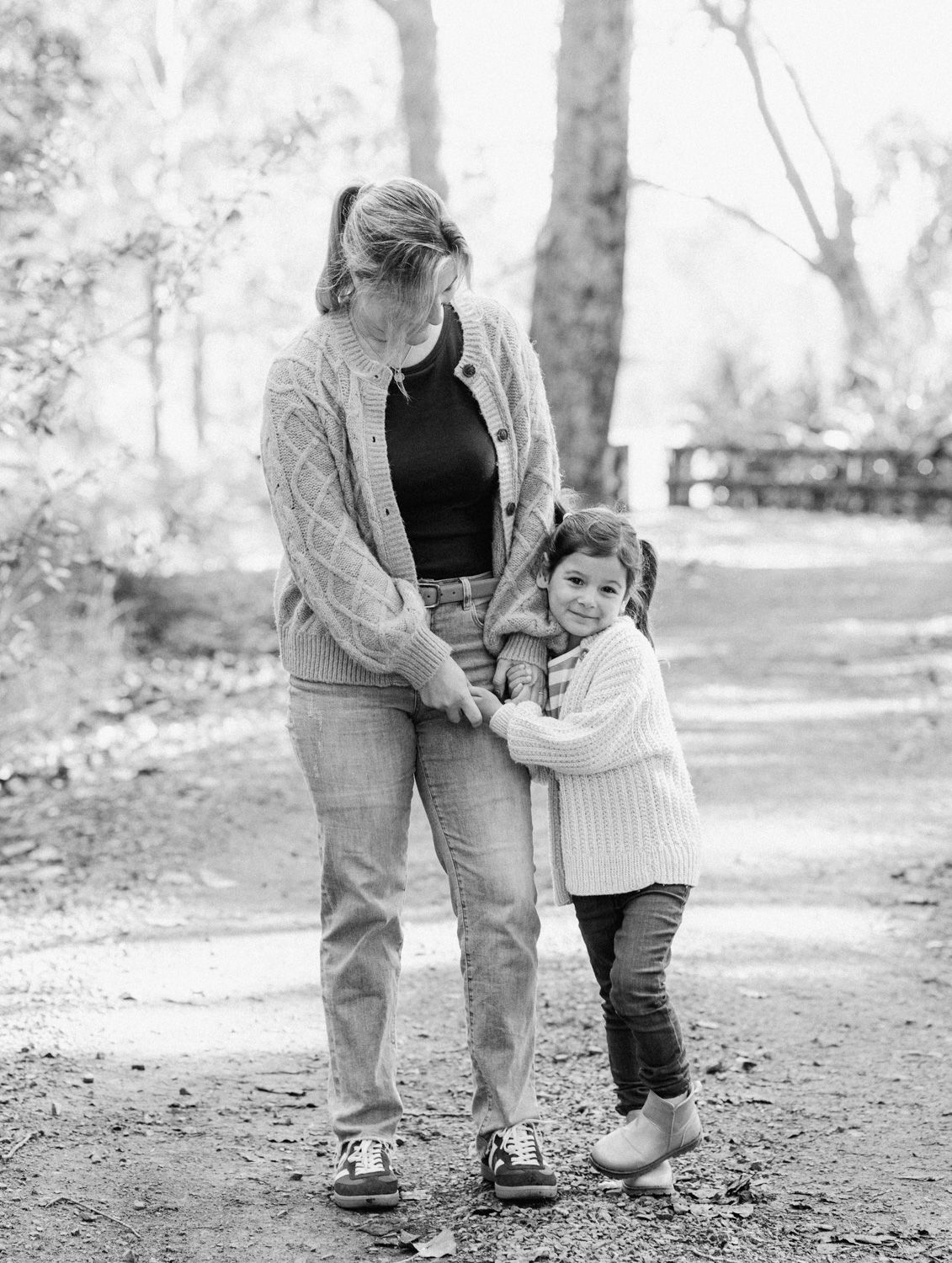 Black and white photo of a mother and young child hugging on a forest path, surrounded by trees.