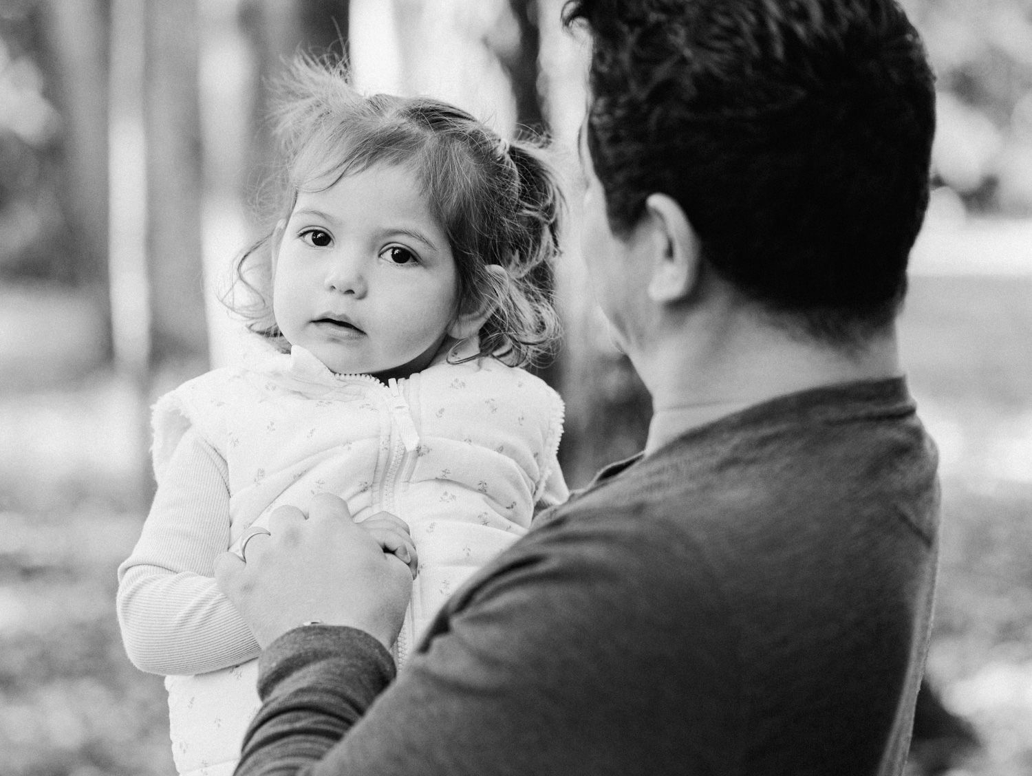 Black and white photo of a father holding his young toddler daughter outdoors among trees.