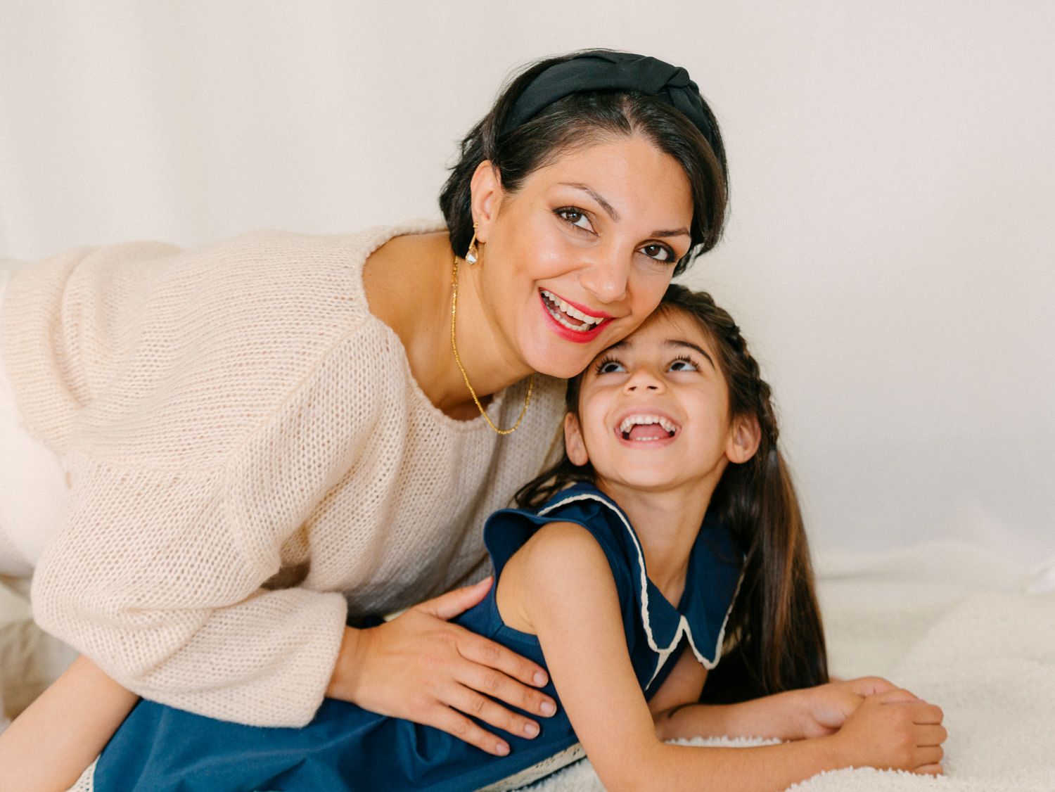 A smiling mother hugs her happy young daughter in a bright, cozy white studio setting.
