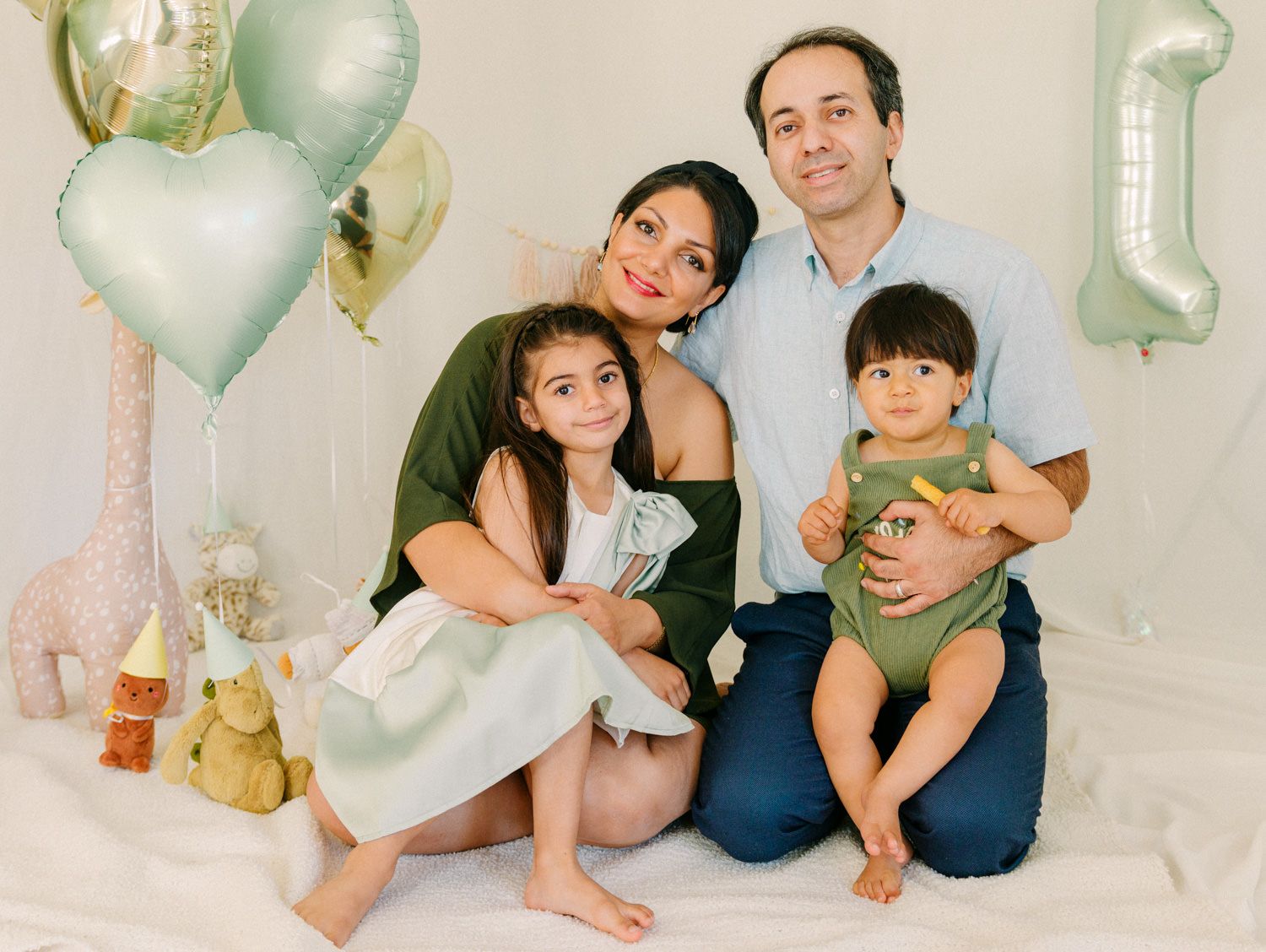 Family of four posing with sage green balloons and toys for a baby's first birthday photo shoot.