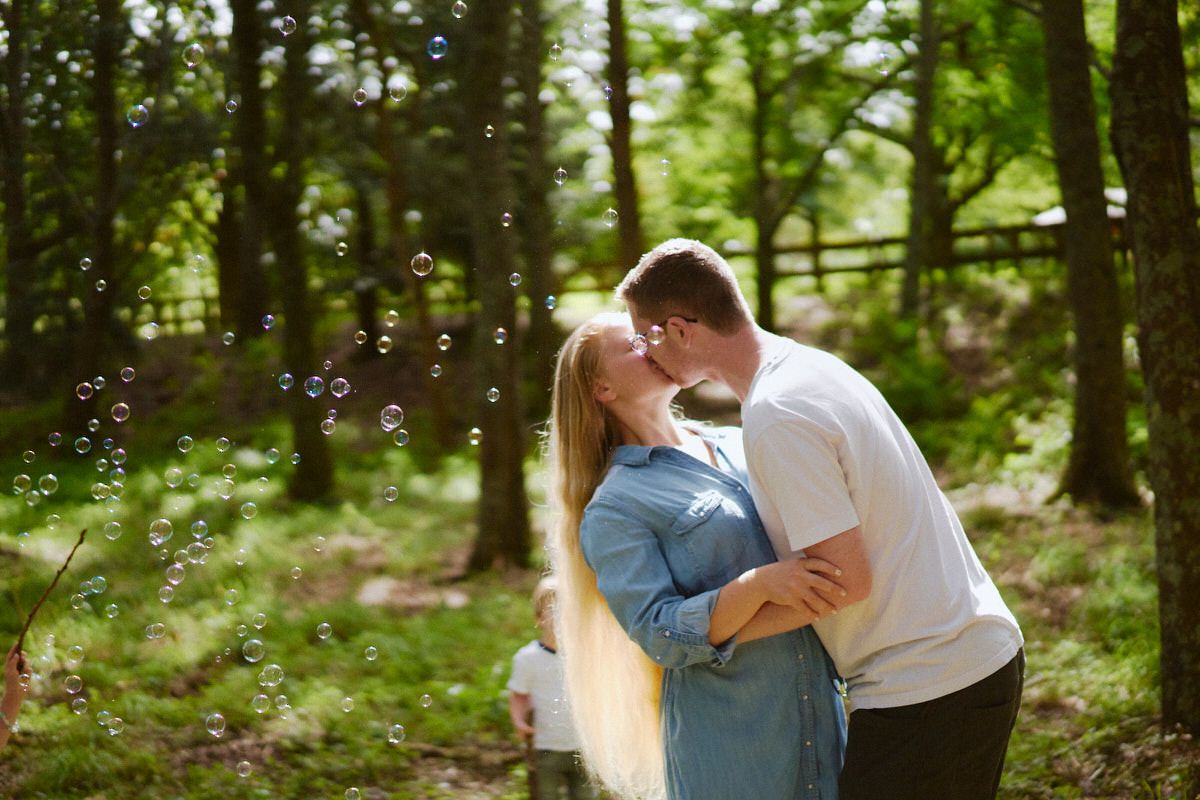 A couple sharing a romantic kiss surrounded by floating bubbles in a sunlit forest.